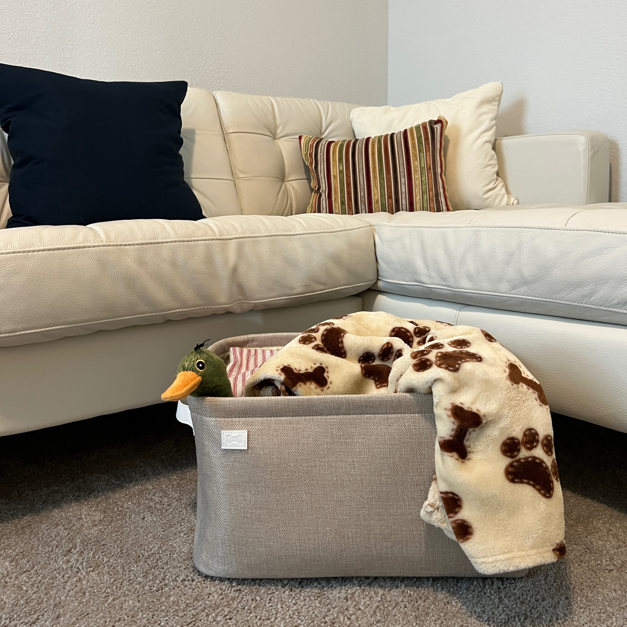 Beige storage bin with blanket and duck toy next to an neutral white couch.