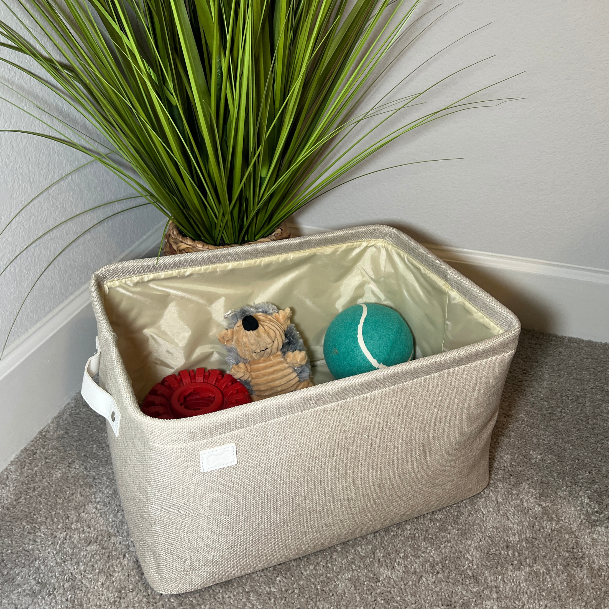 Beige storage bin with toys inside, next to a plant on a carpeted floor.